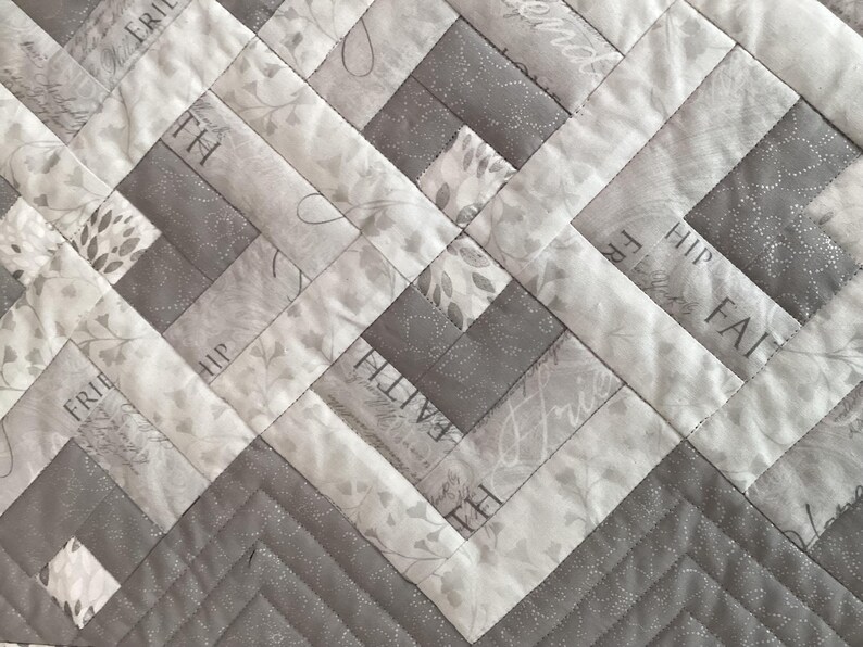 May include: Close-up of a quilted fabric with a geometric design. The quilt features alternating light gray and medium gray rectangular blocks. Some blocks have floral patterns and text that reads "FAITH", "FREE", and "HIP".