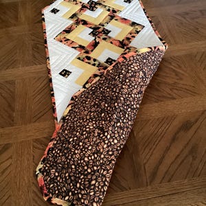 Peut inclure: Chemin de table matelass&eacute; avec un motif g&eacute;om&eacute;trique. Le dessus pr&eacute;sente un tissu blanc, jaune et orange &agrave; motifs. Le dessous est un tissu marron avec un motif de grains de caf&eacute;. Le chemin de table est pos&eacute; sur une table en bois.