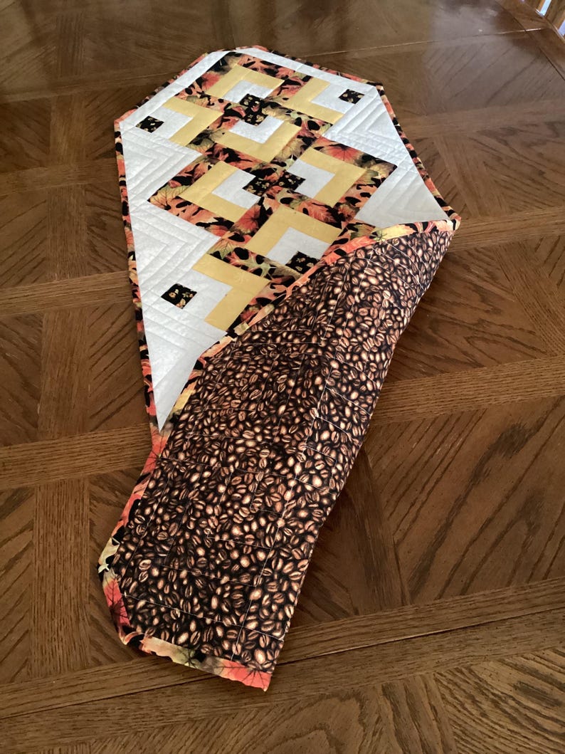 Peut inclure: Chemin de table matelass&eacute; avec un motif g&eacute;om&eacute;trique. Le dessus pr&eacute;sente un tissu blanc, jaune et orange &agrave; motifs. Le dessous est un tissu marron avec un motif de grains de caf&eacute;. Le chemin de table est pos&eacute; sur une table en bois.