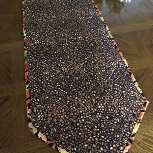 Peut inclure: Chemin de table rectangulaire avec un motif de grains de caf&eacute; sur fond sombre. Les bords sont orn&eacute;s d'un motif floral aux tons de rouge, orange et vert. Pos&eacute; sur une table en bois.