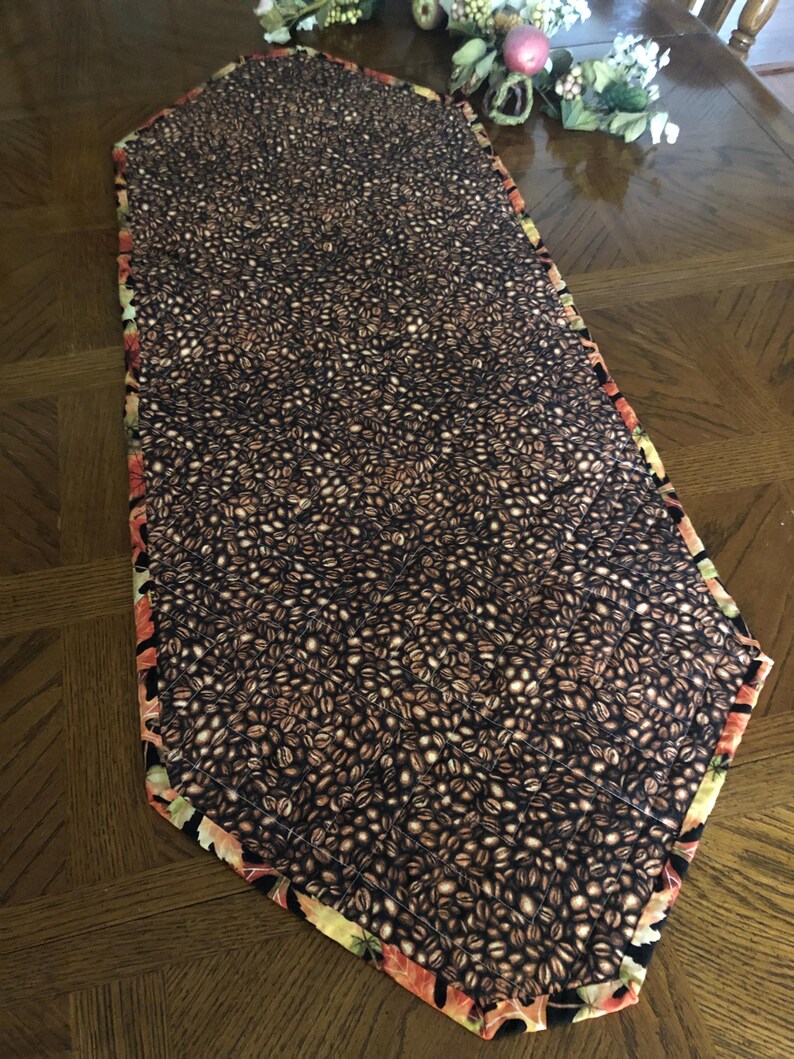 Peut inclure: Chemin de table rectangulaire avec un motif de grains de caf&eacute; sur fond sombre. Les bords sont orn&eacute;s d'un motif floral aux tons de rouge, orange et vert. Pos&eacute; sur une table en bois.