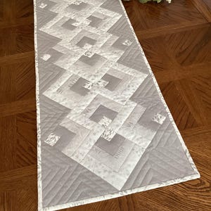 May include: A rectangular gray and white quilted table runner with a geometric design. The runner features a repeating pattern of squares and rectangles, with the words "LOVE", "FAITH", and "SHIP" printed on some of the squares. The edges are finished with a white border.