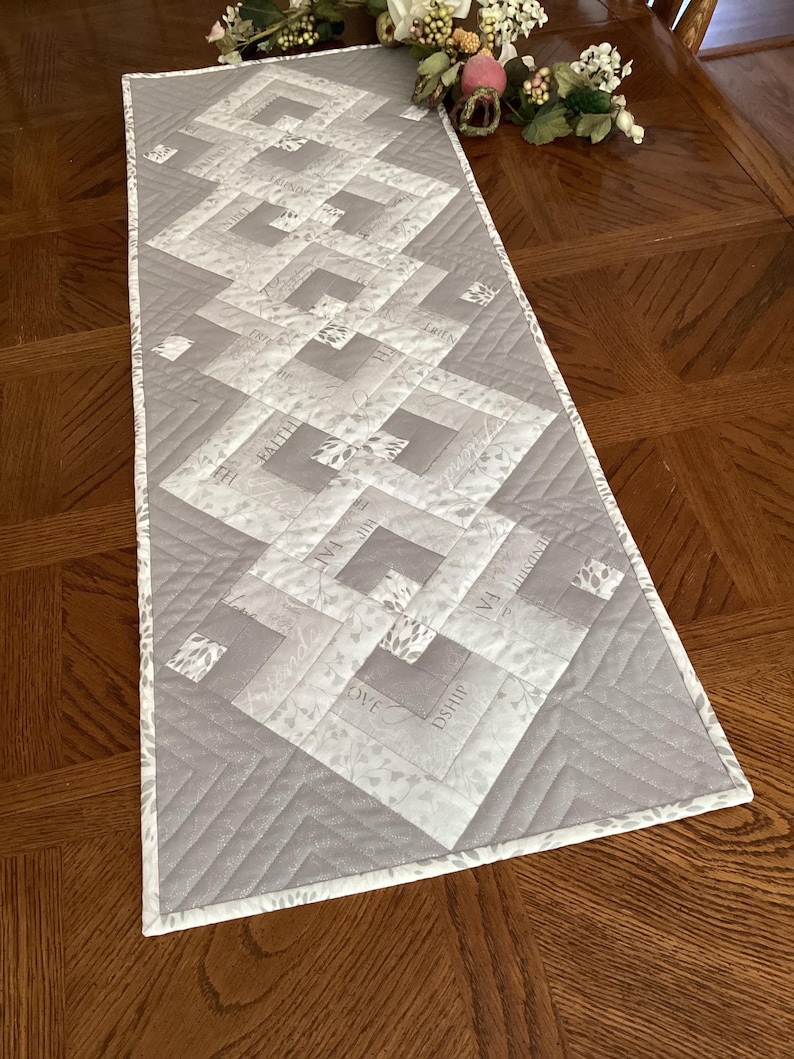 May include: A rectangular gray and white quilted table runner with a geometric design. The runner features a repeating pattern of squares and rectangles, with the words "LOVE", "FAITH", and "SHIP" printed on some of the squares. The edges are finished with a white border.