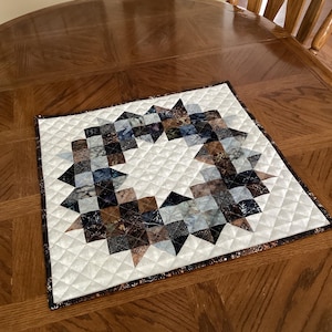 May include: Square quilted placemat with a starburst design. The center features a wreath-like pattern of brown, blue, and black fabric squares. The background is white with a diamond pattern, and the border is a patterned fabric.