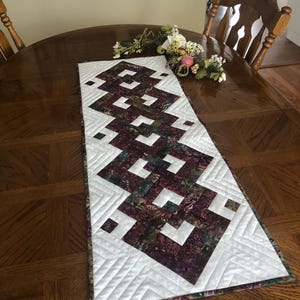 May include: A quilted table runner with a geometric design. The runner features a pattern of interlocking shapes in shades of burgundy, green, and white. The runner is approximately 12 inches wide and 48 inches long.
