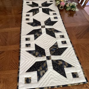 May include: A quilted table runner with a geometric design. The runner features a cream-colored background with black and gold starburst patterns. The edges are finished with a coordinating fabric. The runner is approximately 12 inches wide and 40 inches long.