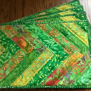 May include: A set of rectangular quilted placemats featuring a diagonal patchwork design. The fabric displays various shades of green, yellow, and orange with floral and leaf patterns. Each placemat is finished with a green checkered border.