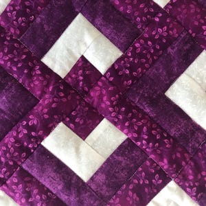 May include: Close-up of a quilted fabric with a geometric pattern. The design features white squares and intersecting bands of purple fabric. The purple fabric has a floral pattern, and the stitching is visible.