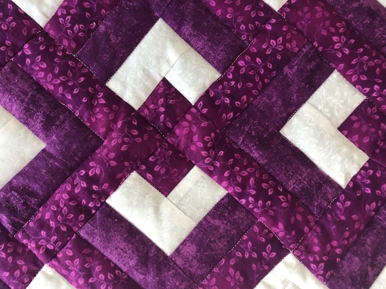 May include: Close-up of a quilted fabric with a geometric pattern. The design features white squares and intersecting bands of purple fabric. The purple fabric has a floral pattern, and the stitching is visible.