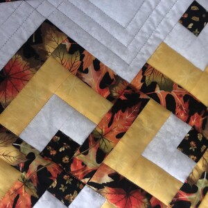 Peut inclure: Gros plan sur une courtepointe avec un motif g&eacute;om&eacute;trique. Le motif pr&eacute;sente du jaune, du blanc, du noir et du tissu avec des motifs de feuilles d'automne. La courtepointe est cousue &agrave; la machine avec un motif r&eacute;p&eacute;titif de lignes obliques.
