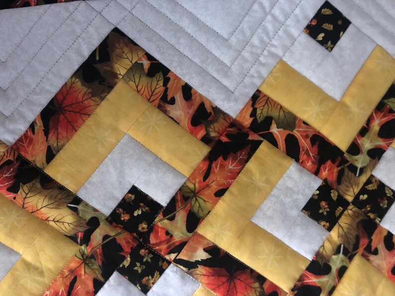 Peut inclure: Gros plan sur une courtepointe avec un motif g&eacute;om&eacute;trique. Le motif pr&eacute;sente du jaune, du blanc, du noir et du tissu avec des motifs de feuilles d'automne. La courtepointe est cousue &agrave; la machine avec un motif r&eacute;p&eacute;titif de lignes obliques.
