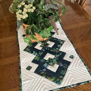 May include: A quilted table runner with a geometric design in shades of navy, green, and white. The runner features a central pattern of interlocking shapes, bordered by a white and green patterned fabric. A basket of faux greenery sits on top.