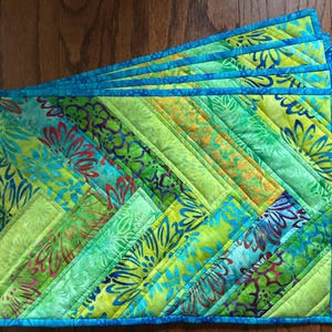 May include: A set of rectangular quilted placemats featuring a vibrant batik pattern in shades of lime green, yellow, and turquoise. The fabric displays a floral motif with diagonal stitching lines and a solid bright blue fabric border trim.