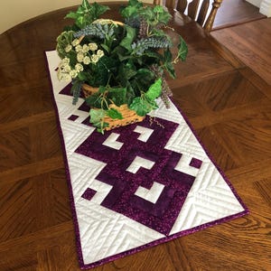 May include: A quilted table runner with a geometric design in white and purple, placed on a wooden table. The runner has a purple border and is topped with a basket of greenery and flowers.