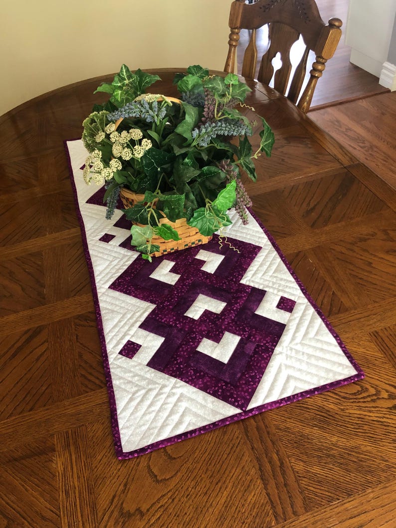 May include: A quilted table runner with a geometric design in white and purple, placed on a wooden table. The runner has a purple border and is topped with a basket of greenery and flowers.