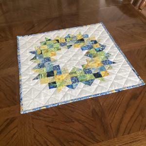 May include: Square quilted placemat with a wreath design. The wreath is made of blue, green, and yellow fabric squares on a white background. The placemat has a blue border and is placed on a wooden table.