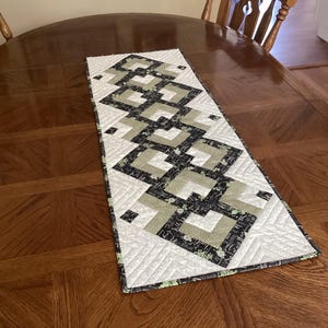 May include: A black, white, and green quilted table runner with a geometric pattern. The runner is on a wooden table.