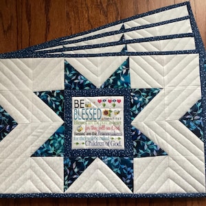 May include: A stack of quilted placemats with a star design. The placemats feature a white center with a blue and white floral pattern and a dark blue border. The center of each placemat has the text "BE BLESSED".