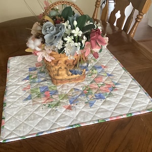 May include: A decorative basket filled with artificial flowers in shades of blue, pink, and white sits on a quilted placemat. The placemat features a geometric pattern in pastel colors on a white background. The basket and placemat are on a wooden table.