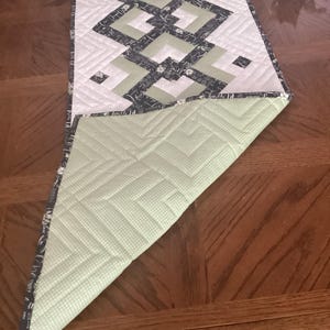 Pode incluir: Um caminho de mesa acolchoado retangular com um padr&atilde;o geom&eacute;trico em preto, branco e verde s&aacute;lvia. A superf&iacute;cie superior apresenta um design quadrado repetitivo, enquanto o canto dobrado revela um tecido verde claro com bolinhas brancas.