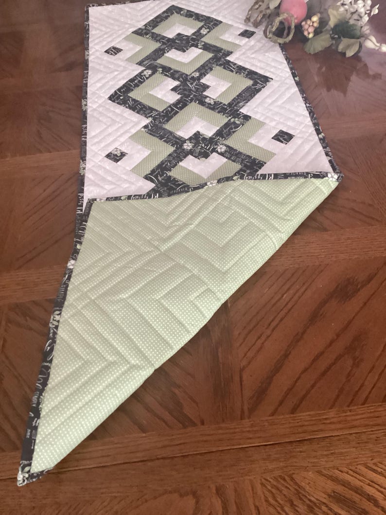 Pode incluir: Um caminho de mesa acolchoado retangular com um padr&atilde;o geom&eacute;trico em preto, branco e verde s&aacute;lvia. A superf&iacute;cie superior apresenta um design quadrado repetitivo, enquanto o canto dobrado revela um tecido verde claro com bolinhas brancas.