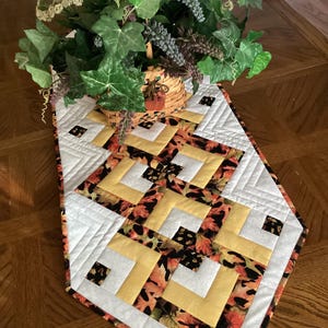 Peut inclure: Chemin de table matelass&eacute; avec un motif g&eacute;om&eacute;trique blanc, jaune et imprim&eacute; de feuilles d'automne. Le chemin de table est pos&eacute; sur une table en bois et surmont&eacute; d'un panier de verdure.