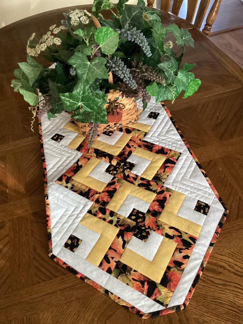 Peut inclure: Chemin de table matelass&eacute; avec un motif g&eacute;om&eacute;trique blanc, jaune et imprim&eacute; de feuilles d'automne. Le chemin de table est pos&eacute; sur une table en bois et surmont&eacute; d'un panier de verdure.