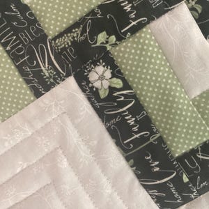 Pode incluir: Uma colcha de retalhos com um padr&atilde;o geom&eacute;trico de tiras de tecido. O design inclui tecido preto com texto branco em cursivo escrito family, love e blessings, tecido verde s&aacute;lvia com bolinhas brancas e tecido off-white com estampas florais.