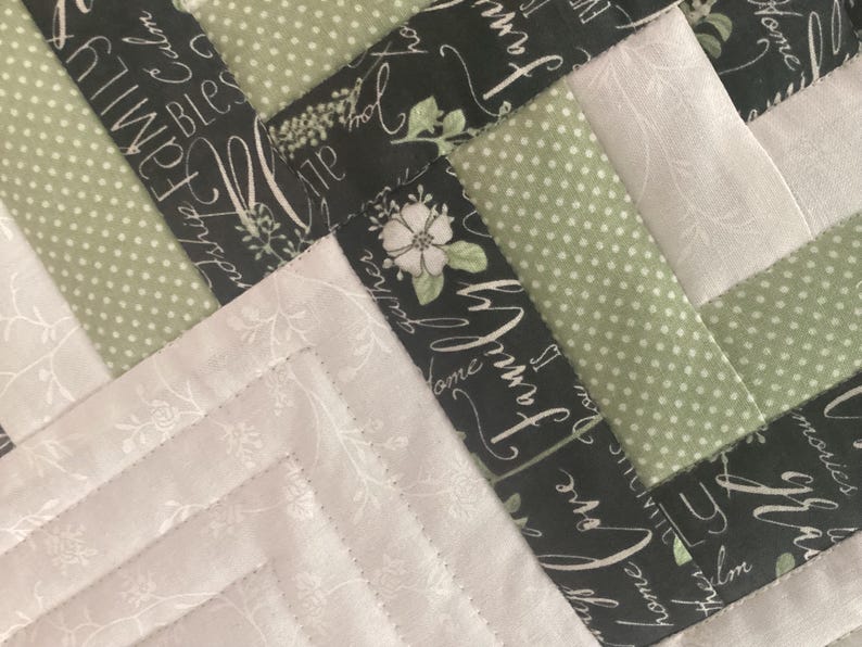 Pode incluir: Uma colcha de retalhos com um padr&atilde;o geom&eacute;trico de tiras de tecido. O design inclui tecido preto com texto branco em cursivo escrito family, love e blessings, tecido verde s&aacute;lvia com bolinhas brancas e tecido off-white com estampas florais.