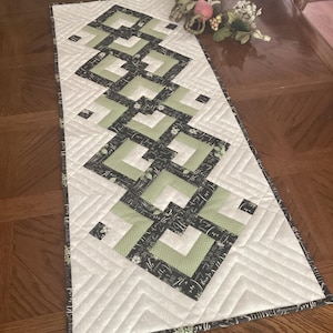 May include: A rectangular quilted table runner featuring a central geometric pattern of sage green and black fabric squares. The runner has a white background with diagonal quilted stitching and a black border printed with white script text.