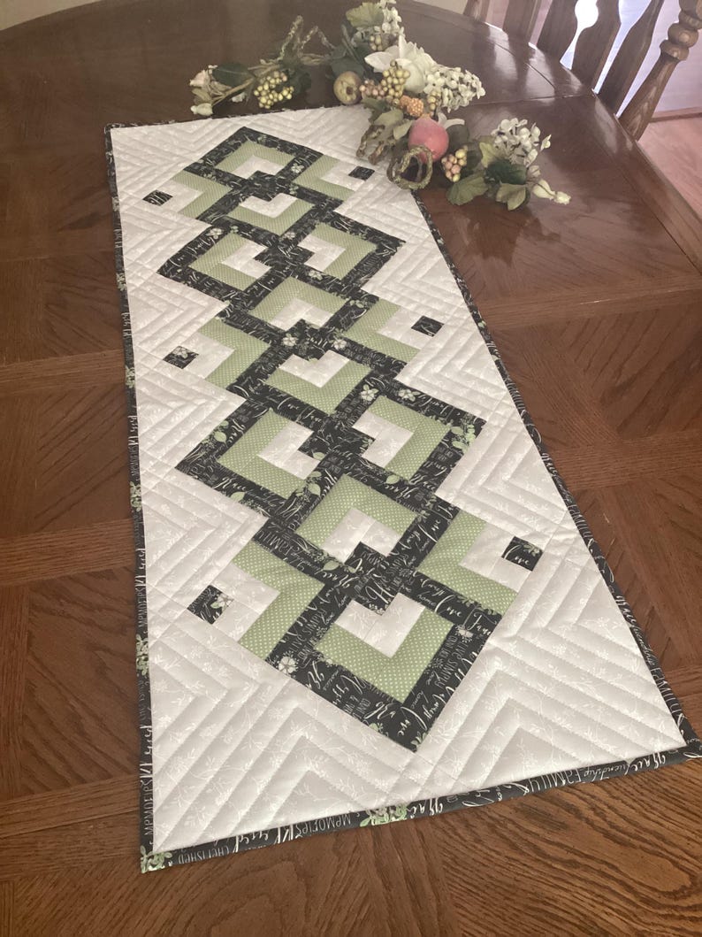 Pode incluir: Um caminho de mesa acolchoado retangular com um padr&atilde;o geom&eacute;trico central de quadrados de tecido verde s&aacute;lvia e preto. O caminho tem um fundo branco com costuras acolchoadas diagonais e uma borda preta estampada com texto branco.