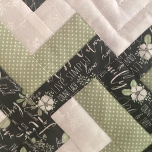 Pode incluir: Uma colcha de retalhos com um padr&atilde;o repetitivo de tecido verde-s&aacute;lvia com bolinhas, tecido branco s&oacute;lido com uma estampa floral sutil e tecido preto estampado com flores brancas e o texto "Live simply, count each blessing."