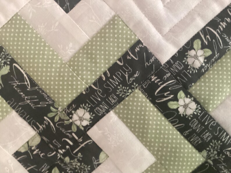 Pode incluir: Uma colcha de retalhos com um padr&atilde;o repetitivo de tecido verde-s&aacute;lvia com bolinhas, tecido branco s&oacute;lido com uma estampa floral sutil e tecido preto estampado com flores brancas e o texto "Live simply, count each blessing."