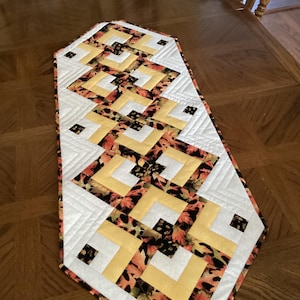 May include: A quilted table runner with a geometric design. The runner features a white border, yellow squares, and a leaf pattern in orange, brown, and black. The runner is placed on a wooden table.