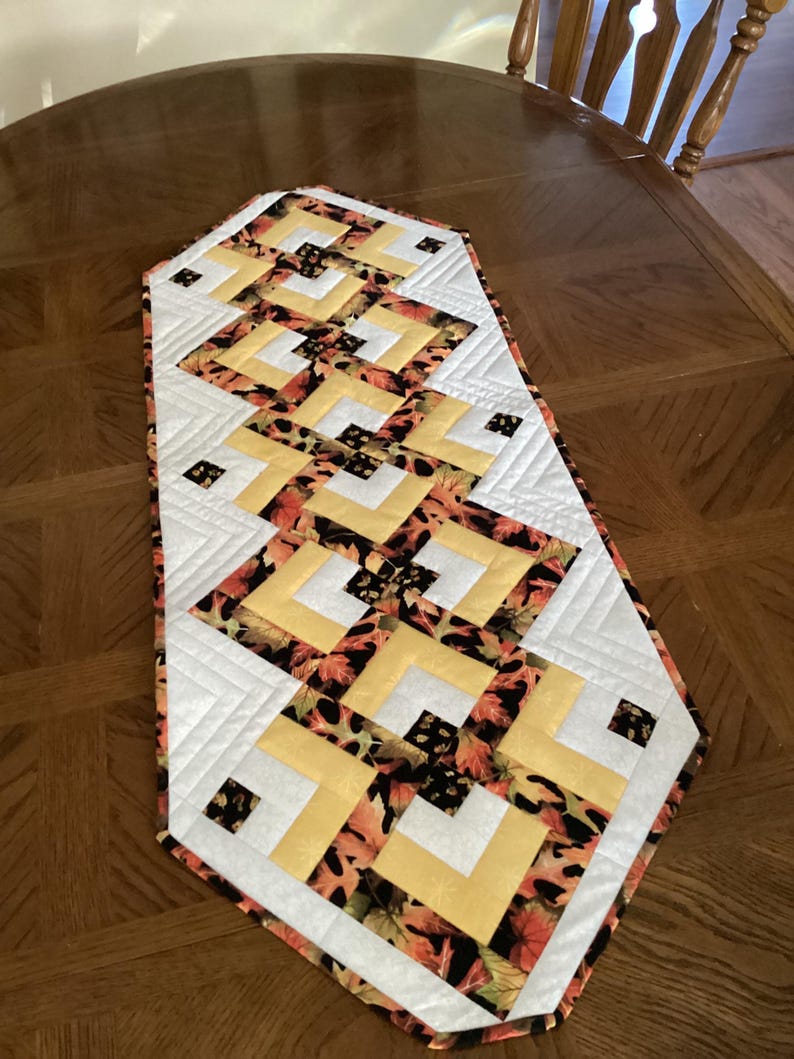 Peut inclure: Chemin de table matelass&eacute; avec un motif g&eacute;om&eacute;trique. Le chemin de table pr&eacute;sente une bordure blanche, des carr&eacute;s jaunes et un motif de feuilles orange, marron et noir. Pos&eacute; sur une table en bois.