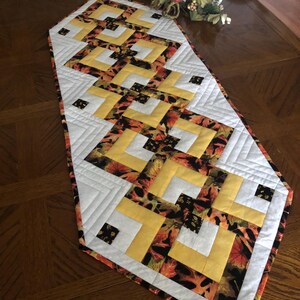 Peut inclure: Chemin de table matelass&eacute; avec un motif g&eacute;om&eacute;trique. Le chemin de table pr&eacute;sente un motif de tissu &agrave; imprim&eacute; feuilles jaune, blanc et aux couleurs automnales. Le chemin de table est pos&eacute; sur une table en bois, mettant en valeur son attrait d&eacute;coratif.