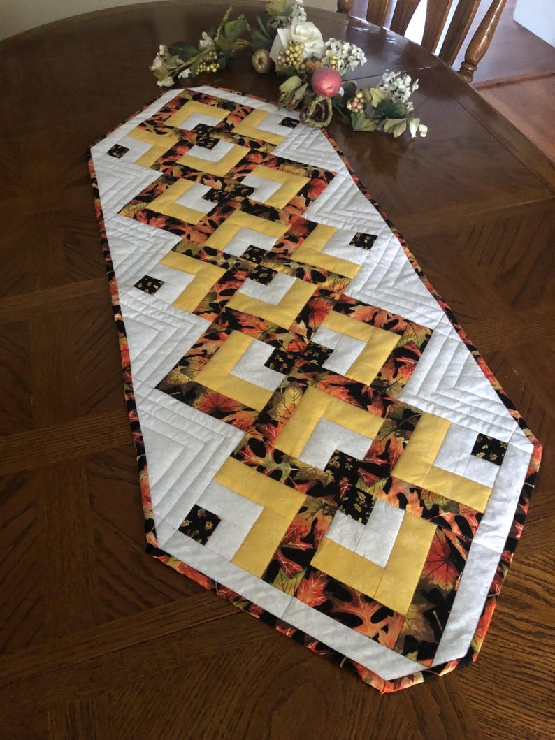Peut inclure: Chemin de table matelass&eacute; avec un motif g&eacute;om&eacute;trique. Le chemin de table pr&eacute;sente un motif de tissu &agrave; imprim&eacute; feuilles jaune, blanc et aux couleurs automnales. Le chemin de table est pos&eacute; sur une table en bois, mettant en valeur son attrait d&eacute;coratif.