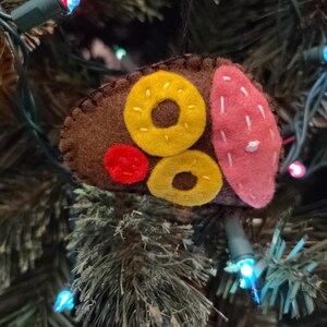 May include: A felt Christmas ornament shaped like a sushi roll. The ornament features a brown base with yellow donut-shaped pieces, a red circle, and a pink piece. The ornament is hanging on a Christmas tree with colorful lights.