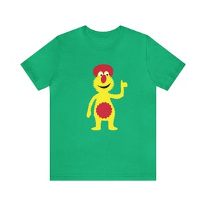 May include: A green t-shirt featuring a yellow cartoon character wearing a red hat and a red circle on its chest. The character is giving a thumbs up.