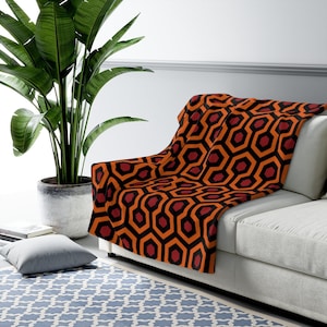 May include: A red and orange throw blanket with a geometric pattern. The blanket is folded and draped over a white couch.