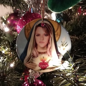 May include: A round Christmas tree ornament featuring a portrait of a woman with a halo and a blue veil. The ornament has a gold border and a red heart with flames. The ornament is hanging on a Christmas tree.
