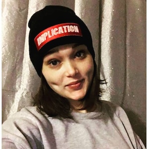 May include: A black beanie with a red patch that says "IMPLICATION" in white letters.