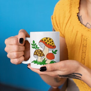 Peut inclure: Une tasse en c&eacute;ramique blanche avec un motif de champignons color&eacute;s. Les champignons sont rouges, jaunes, bruns et verts. La tasse a une poign&eacute;e noire.