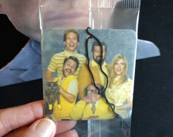 May include: A car air freshener featuring a group of people from the TV show 'It's Always Sunny in Philadelphia'. The characters are depicted in a yellow, brown, and blue color scheme. The air freshener is packaged in a clear plastic bag with a black string.