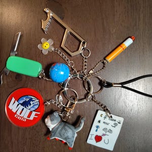 May include: A collection of keychains and charms, including a green pocket knife, a blue globe, a red Wolf Cola logo, a cat figurine, and a small white card with the words "I ❤️ U". Other charms include a flower, a cigarette, and a bottle opener.
