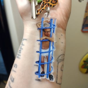 May include: A keychain featuring a clear plastic charm with a photo of a man in a blue cage. The man is wearing white underwear and has glasses. The keychain is attached to a set of keys with a leopard print key chain.