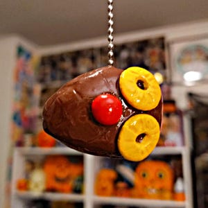 May include: A brown, glazed ham-shaped ornament hanging from a silver ball chain. The ornament features two yellow pineapple rings and a single bright red cherry attached to the top surface, mimicking a classic glazed ham presentation.