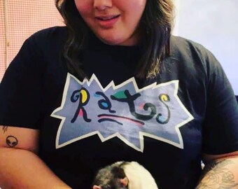 May include: Black t-shirt with the word "Rats" in a colourful, cartoon-style font. The shirt is worn by a person holding a black and white rat. The rat is the focus of the image.