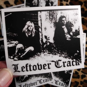 May include: Black and white sticker featuring two figures and the band name "Leftover Crack" in gothic font. The sticker has a rectangular shape with a white border. The background is a leopard print.