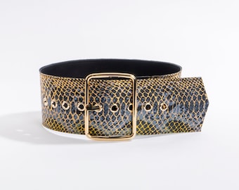 May include: A wide belt featuring a yellow and black snakeskin pattern with a large, rectangular, polished gold-tone metal buckle. The belt includes a series of evenly spaced gold-tone metal eyelets along the length of the strap.
